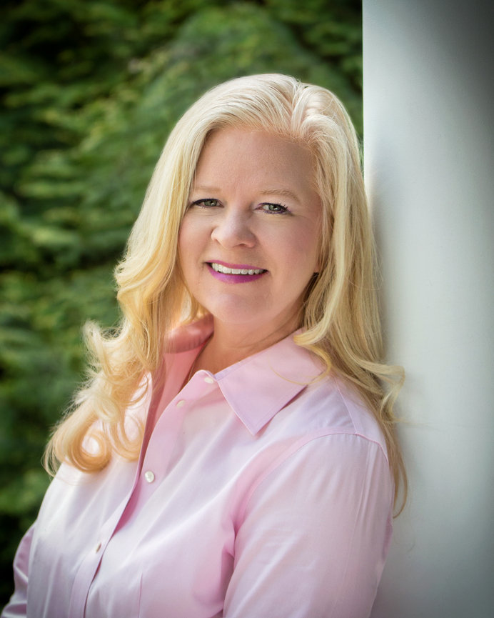 A woman with long blonde hair wearing a pink shirt leans against a white pillar, with greenery in the background.