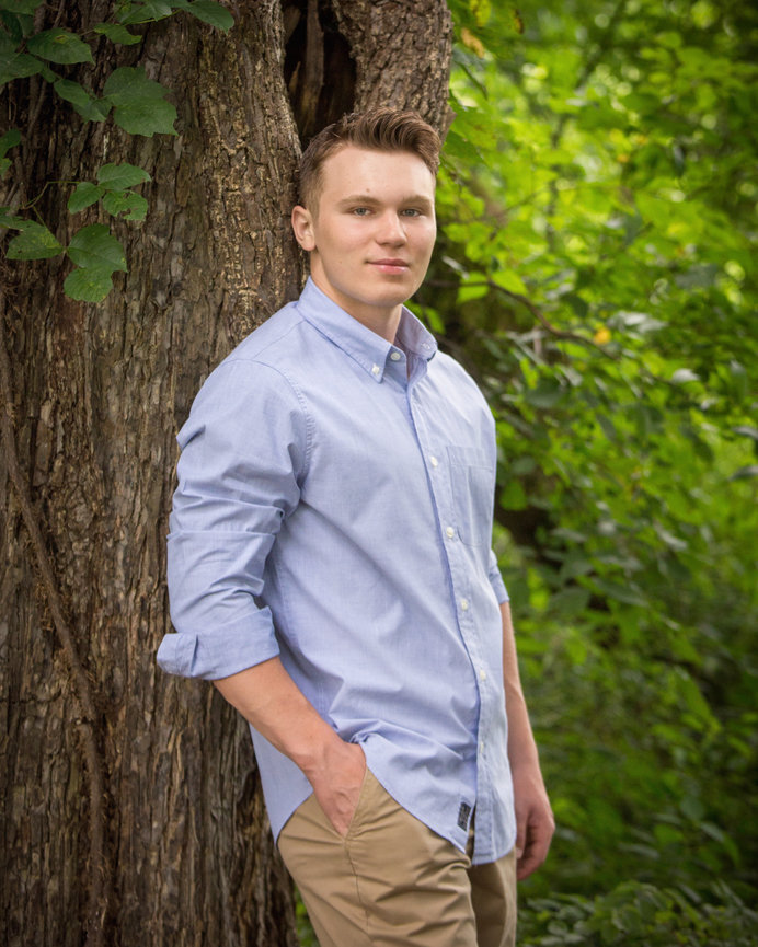 Man in a blue shirt leaning against a tree in a lush green forest setting.