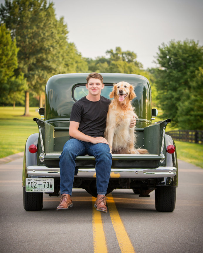 Young man with a golden retriever sitting on a vintage truck's tailgate, parked on a tree-lined road.