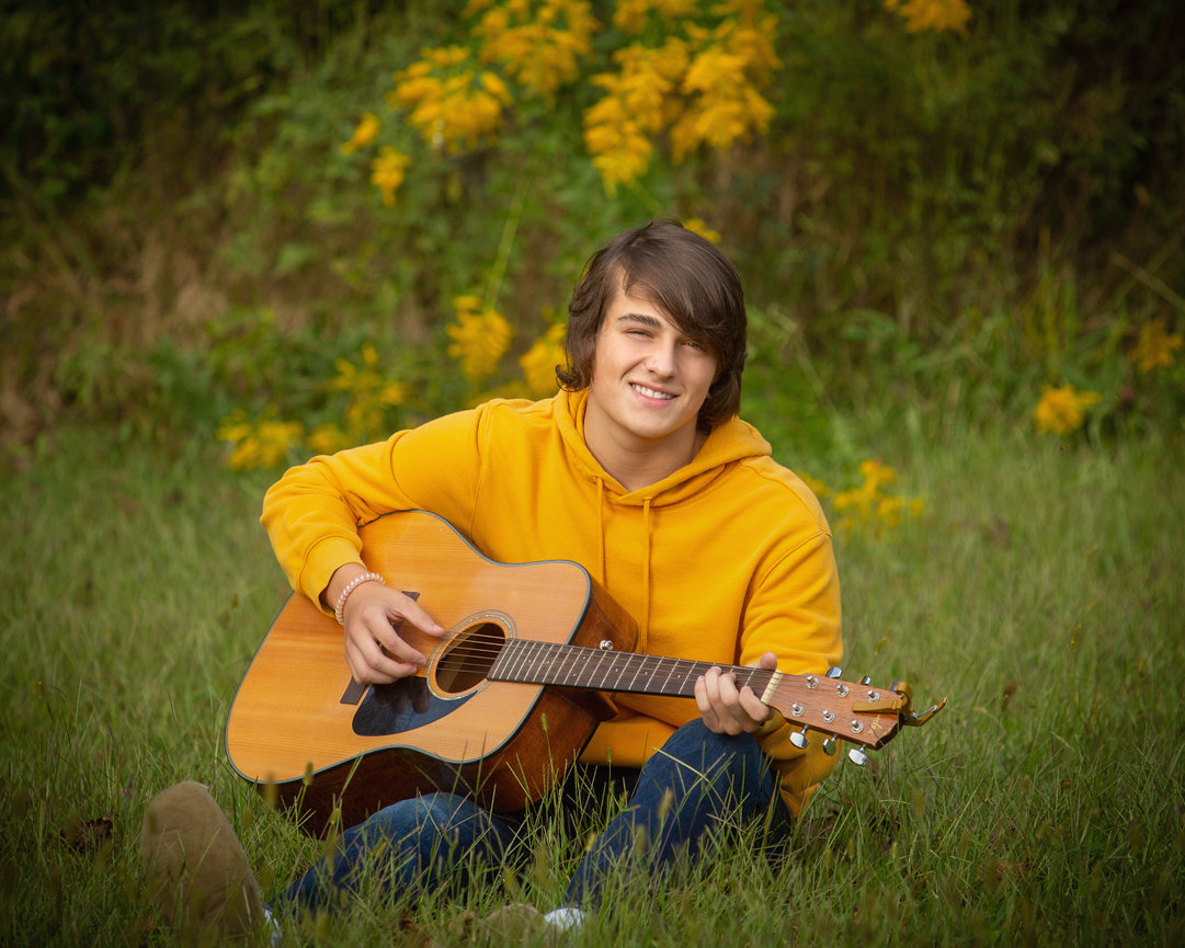 Teen in yellow hoodie playing acoustic guitar while sitting on grass with yellow flowers in background.
