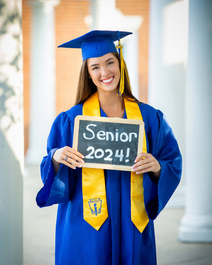 A smiling graduate in a blue cap and gown holds a sign reading Senior 2024 outdoors.