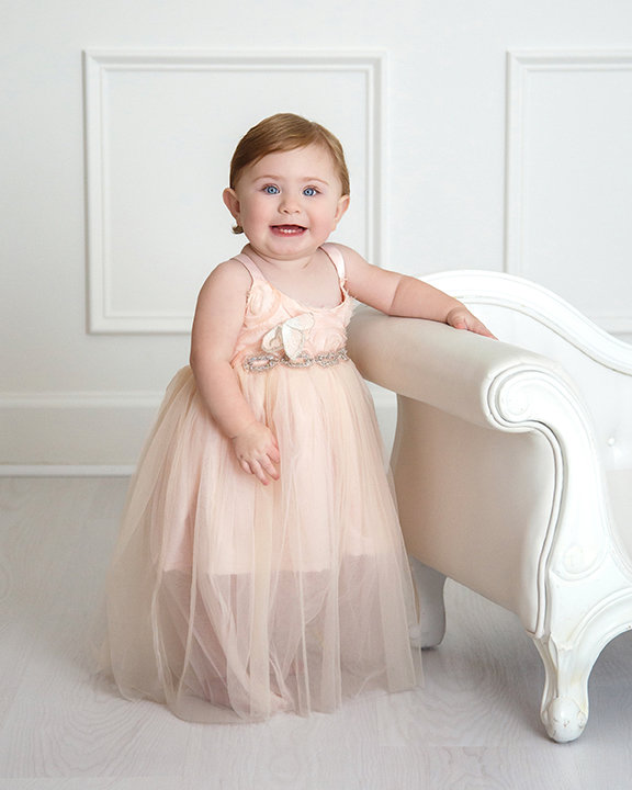 A toddler in a pink dress smiles while standing beside a white chair in a bright room.