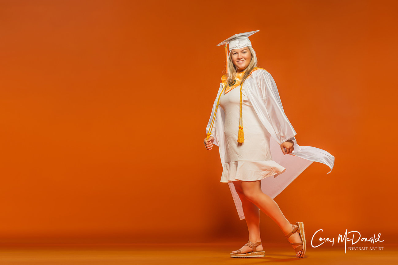 Graduate in a white cap and gown smiling against an orange background.