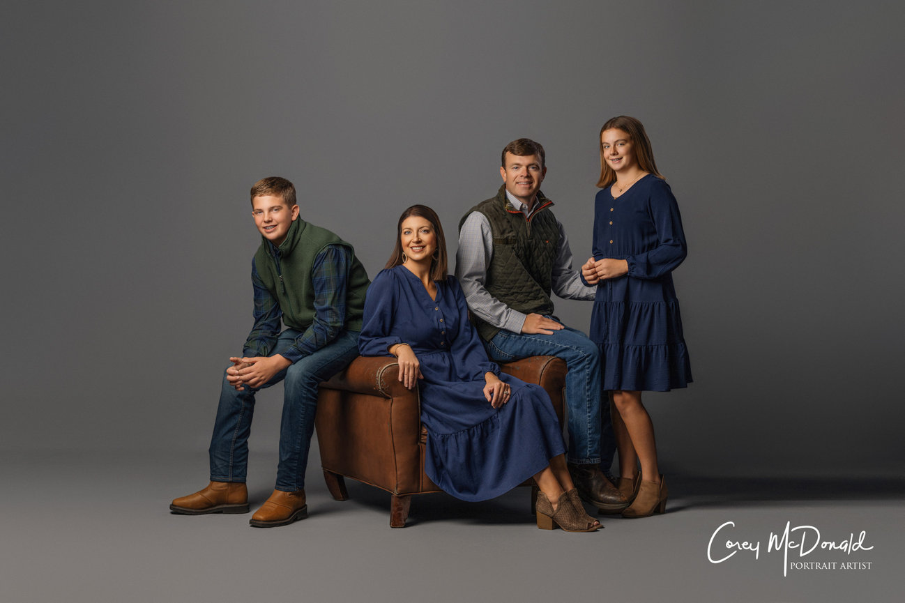 A family of four in blue and neutral outfits poses together on a brown chair against a gray backdrop.