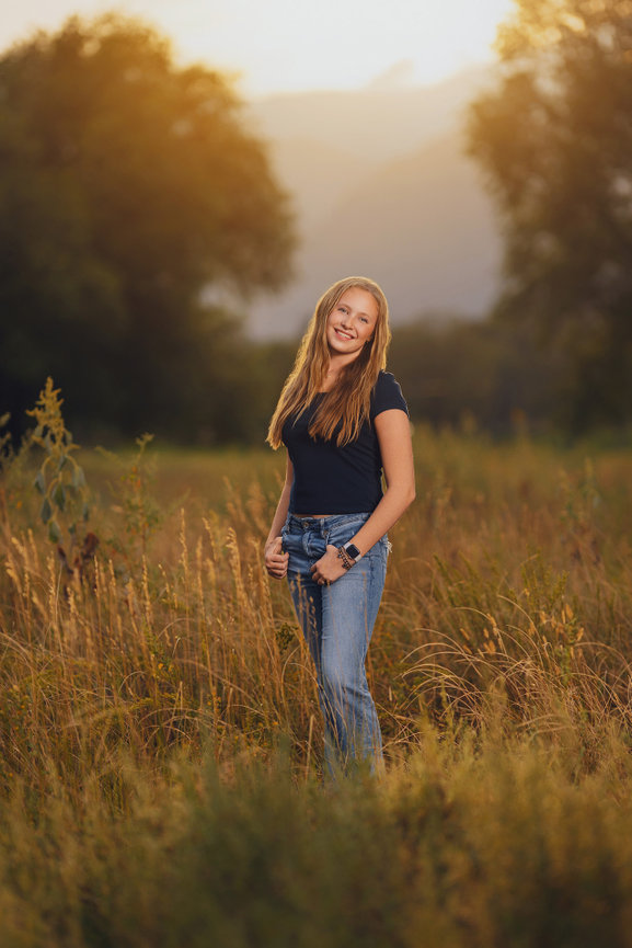 Smiling person standing in a sunlit field with tall grass and trees in the background.