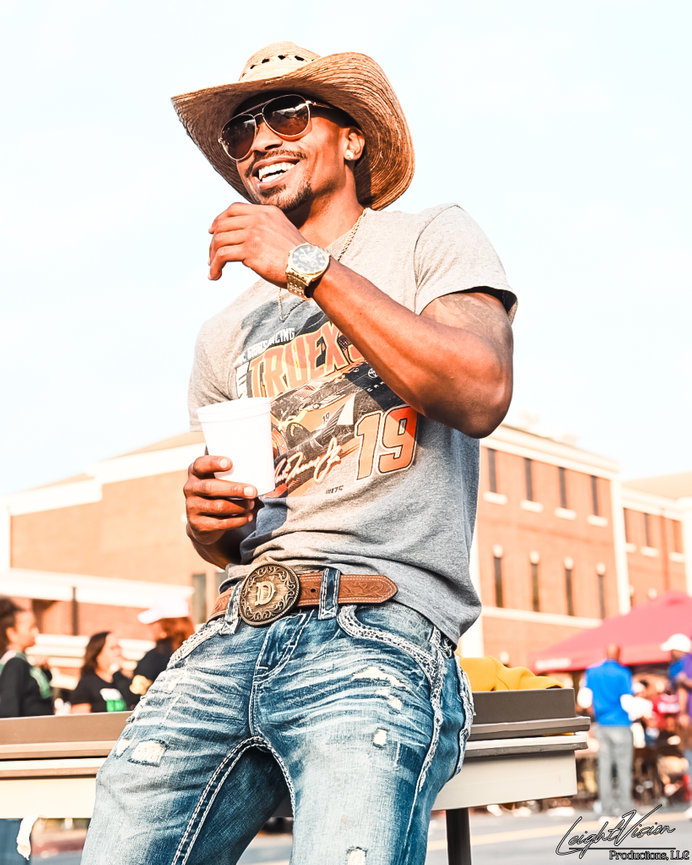 A cheerful man in a cowboy hat and jeans, smiling warmly at the camera.