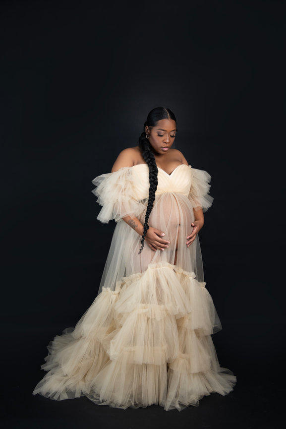 Pregnant woman in a flowing, off-shoulder tulle gown cradling her belly against a dark background.