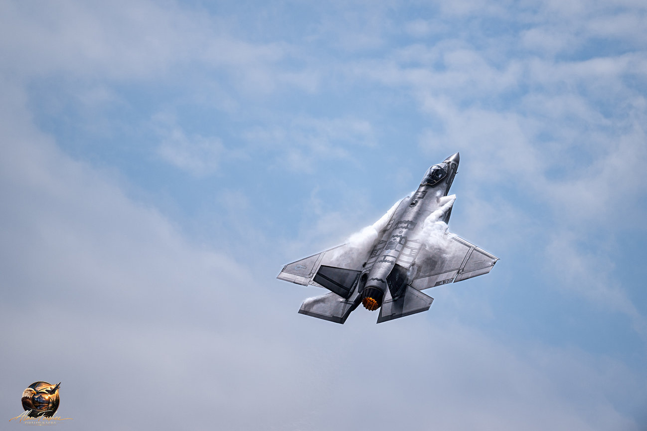 U.S. Air Force F-35 Lightening II climbing with afterburner glowing and vapor forming over its wings against a cloudy blue sky.