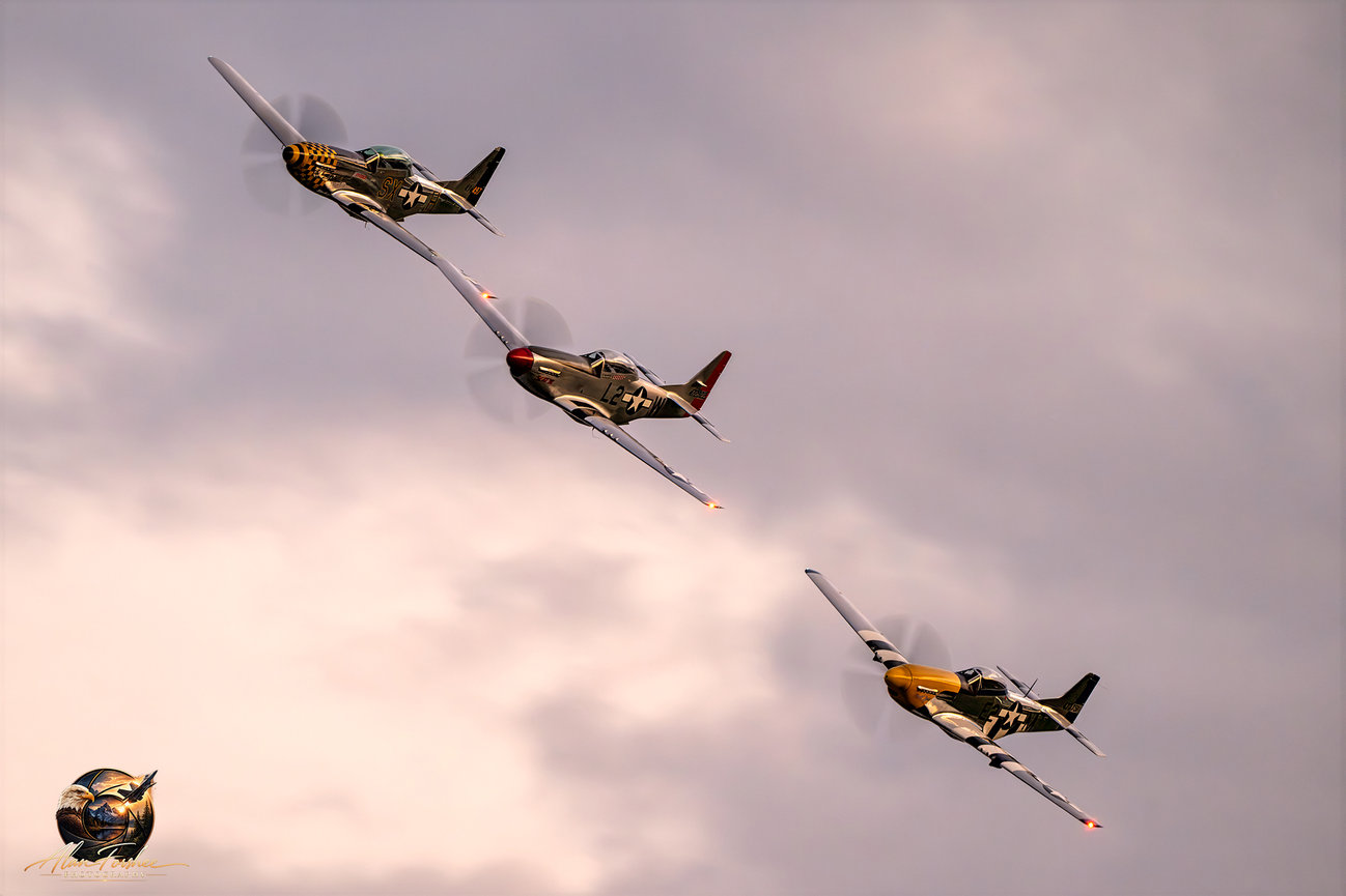 Three vintage P-51 Mustang fighter aircraft flying in formation against a softly illuminated pink and gray evening sky.