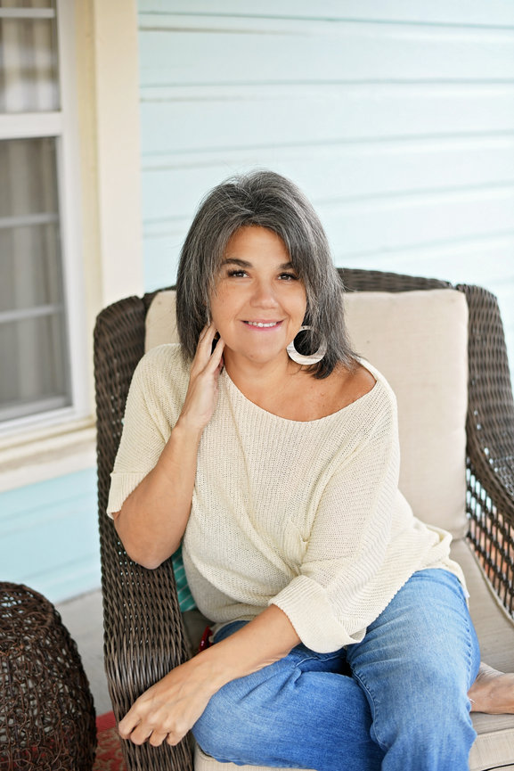 Woman with gray hair sitting on porch chair wearing cream top and jeans.
