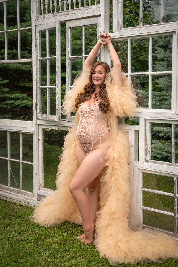 Woman in a floral corset and flowing beige robe poses joyfully against a backdrop of rustic window frames outdoors.