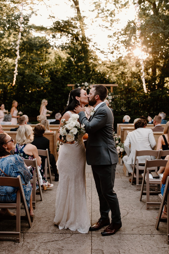 A couple kissing during their outdoor wedding ceremony, surrounded by guests seated on wooden chairs.