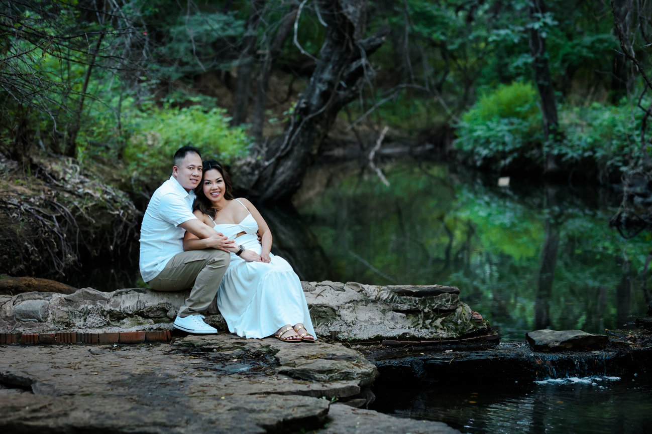 Couple in white outfits sitting on a rock by a forest stream, surrounded by lush greenery.