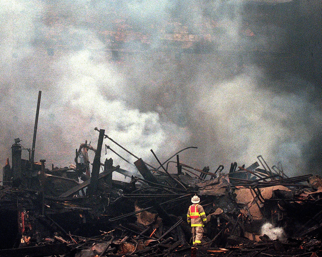 Firefighter amid smoky ruins after a large fire, with debris and thick smoke filling the scene.