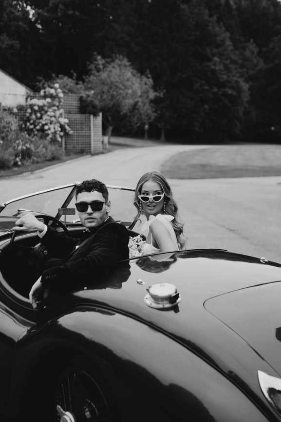 A relaxed, editorial wedding moment at Standon Hall: bride and groom in a classic convertible on the tree-lined drive of The Salt House.