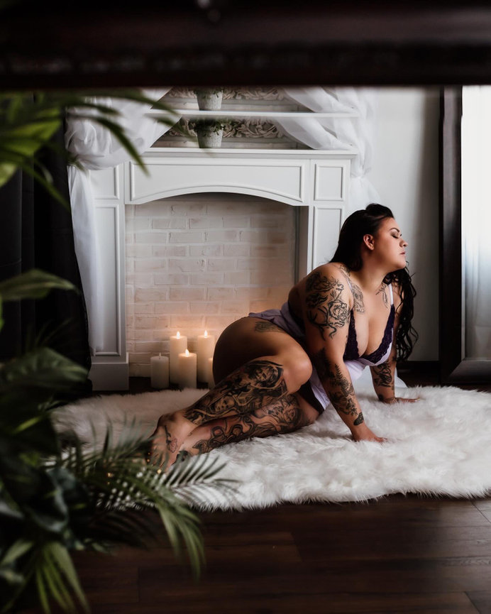 Amy matney boudoir studio port angeles Woman with tattoos poses on a fluffy white rug by a fireplace, surrounded by lit candles and plants.