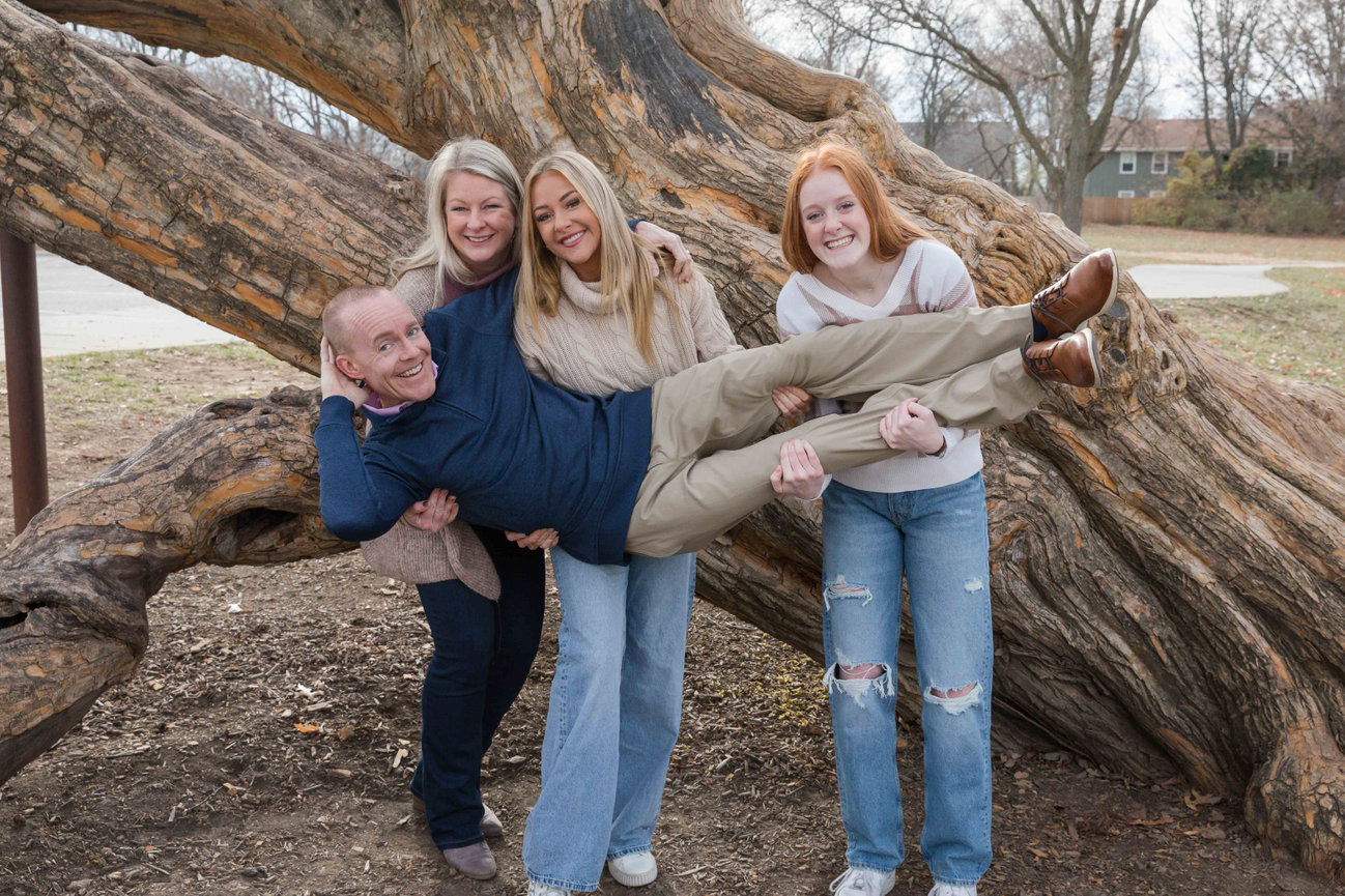 Family of four posing by a large fallen tree, with a man playfully held horizontally