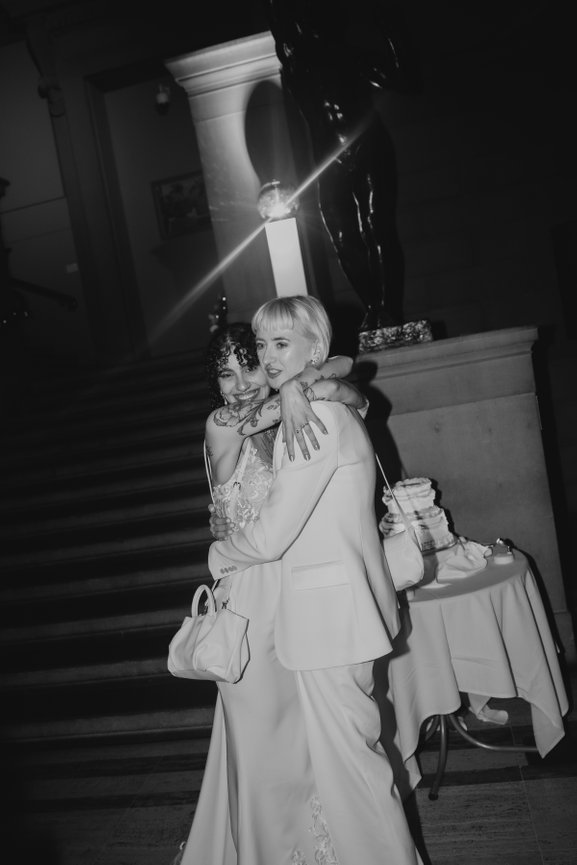 Modern editorial wedding photography in Manchester Art gallery, of two brides embracing and smiling by their wedding cake, captured with direct flash.