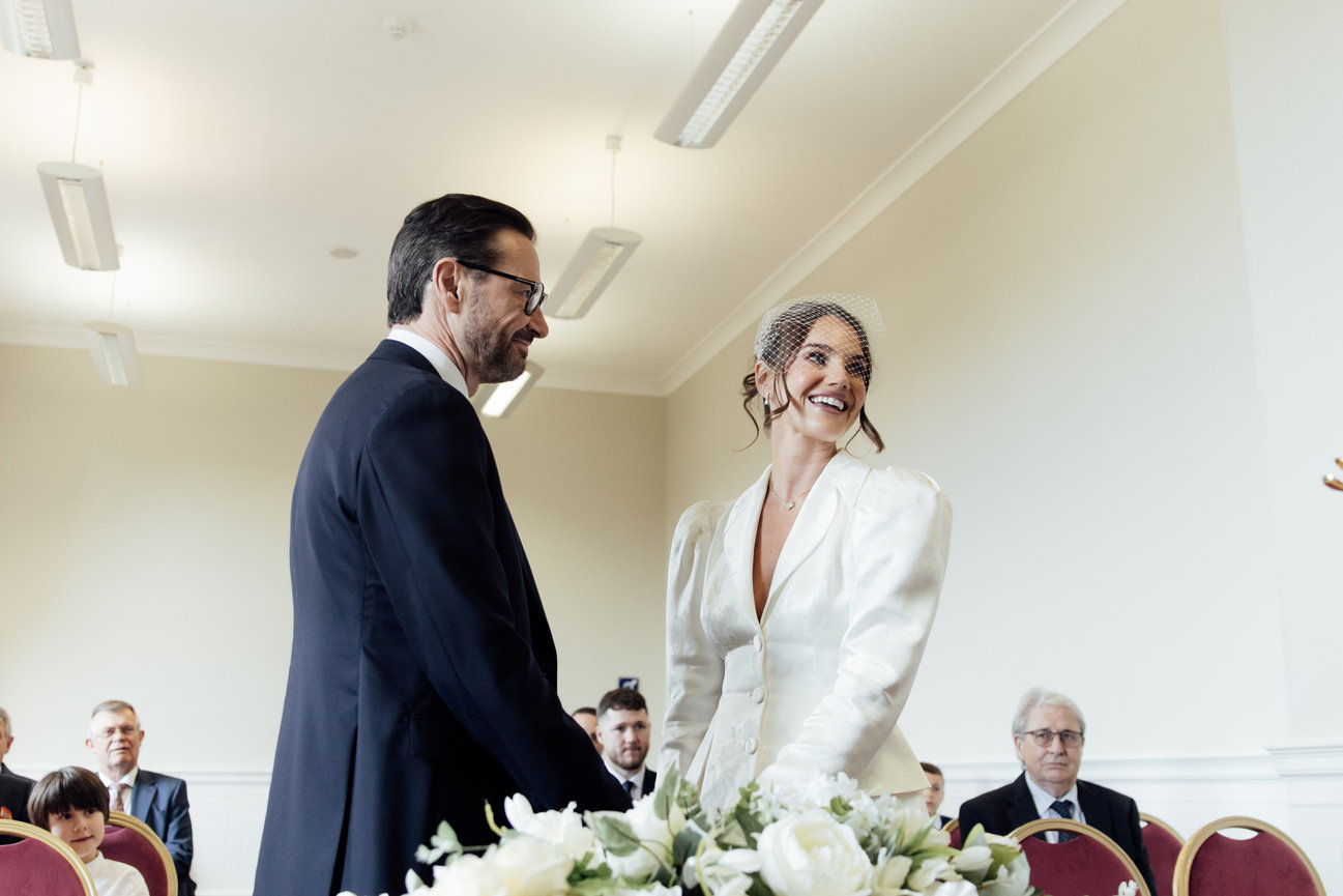 Modern wedding photography: Bride and groom smiling at altar in a room in Liverpool with guests, bouquet in foreground.