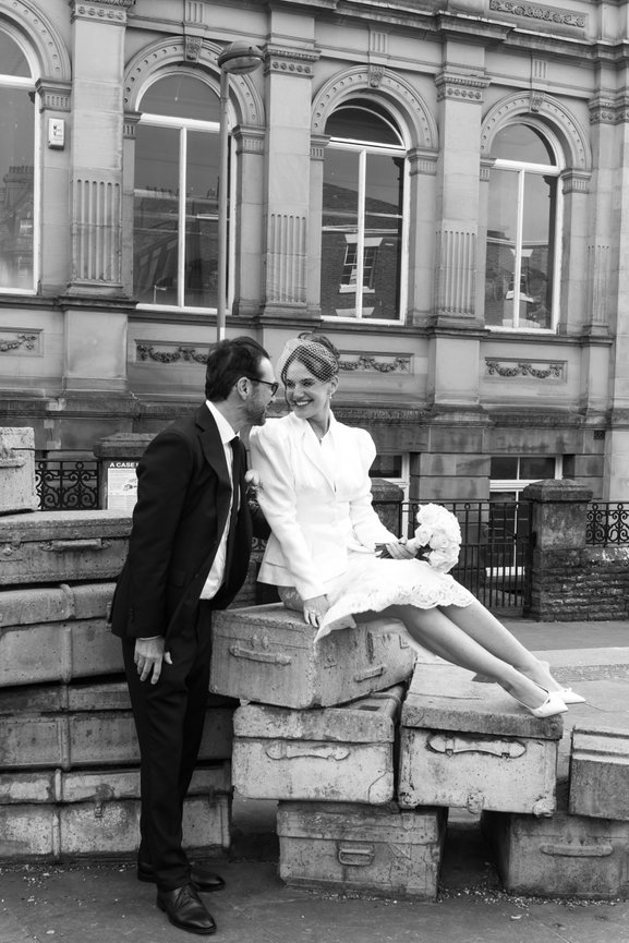 Relaxed editorial wedding photography of a bride sitting on the Hope Street suitcases with a white rose bouquet, smiling at her groom in front of the Liverpool Institute for Performing Arts.