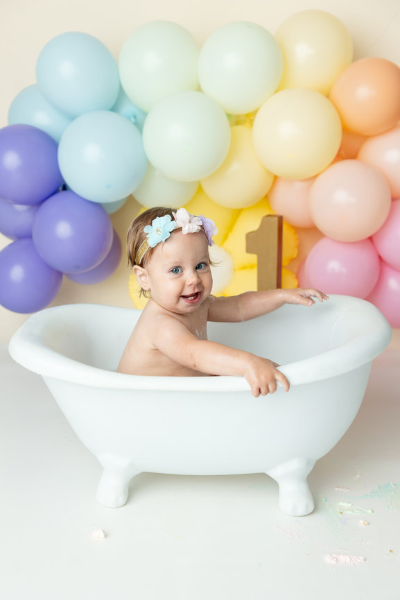 Baby in a white tub with rainbow balloons and a number one sign in the background.