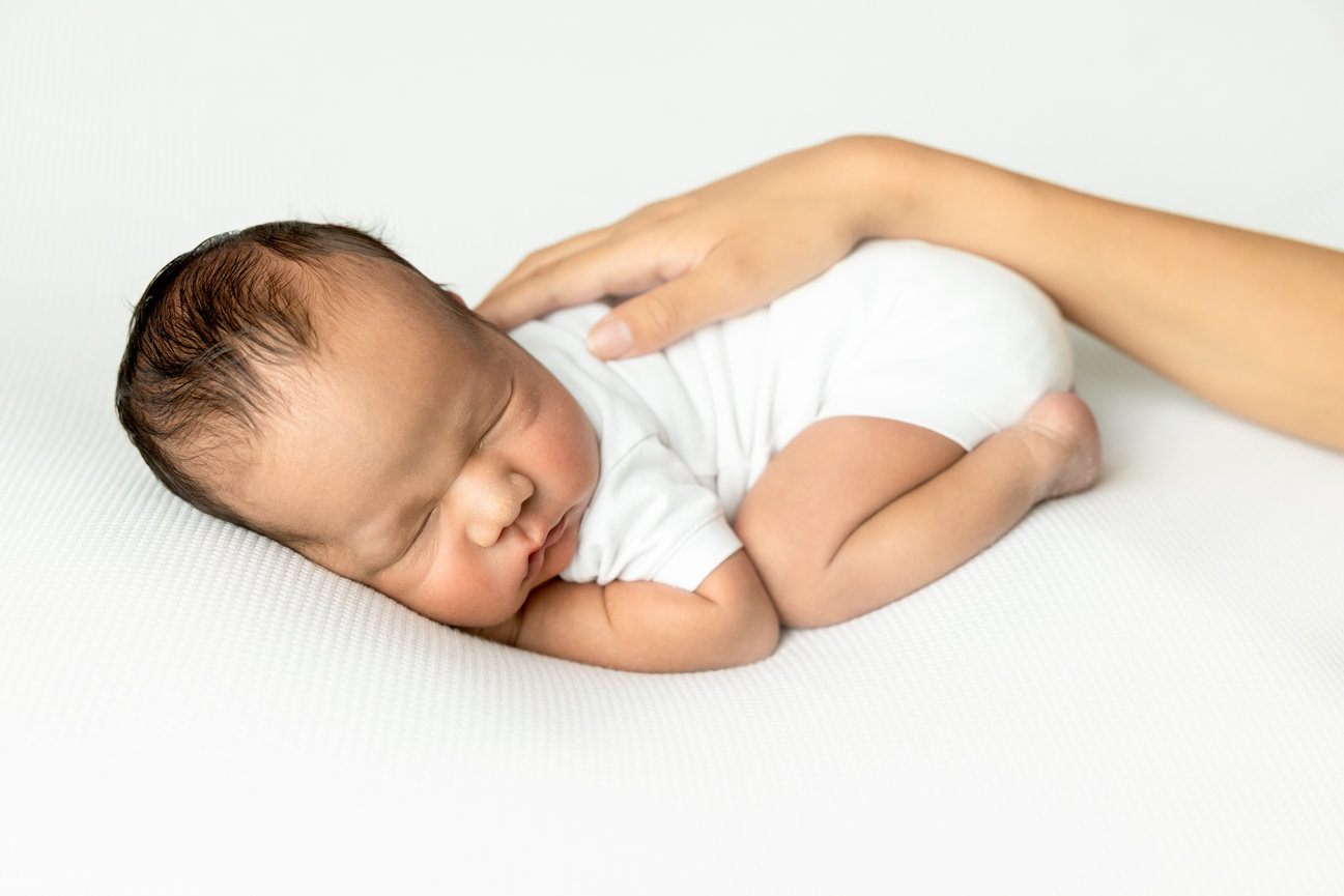 Sleeping newborn in white onesie on a white background with a hand gently resting on their back.