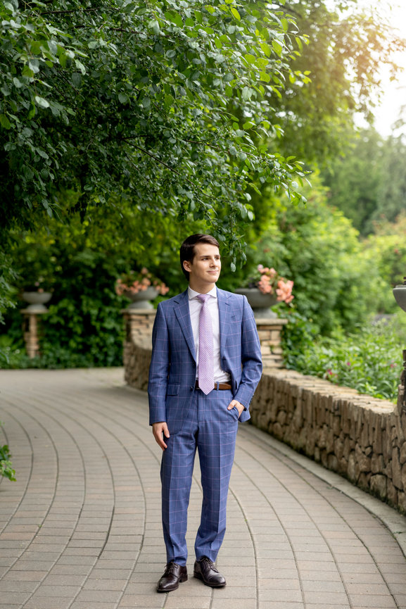 Graduate in a blue suit standing on a garden path under leafy trees in soft sunlight