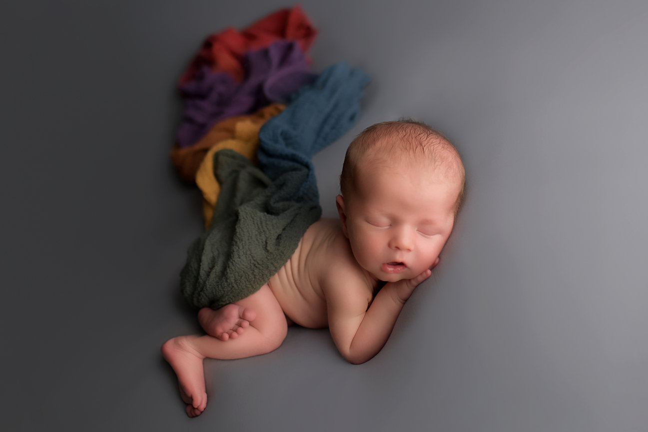 Newborn baby sleeping in bum up pose on grey fabric with rainbow wrap covering him at newborn photography session with Kansas City newborn photographer Dreamworks Photography