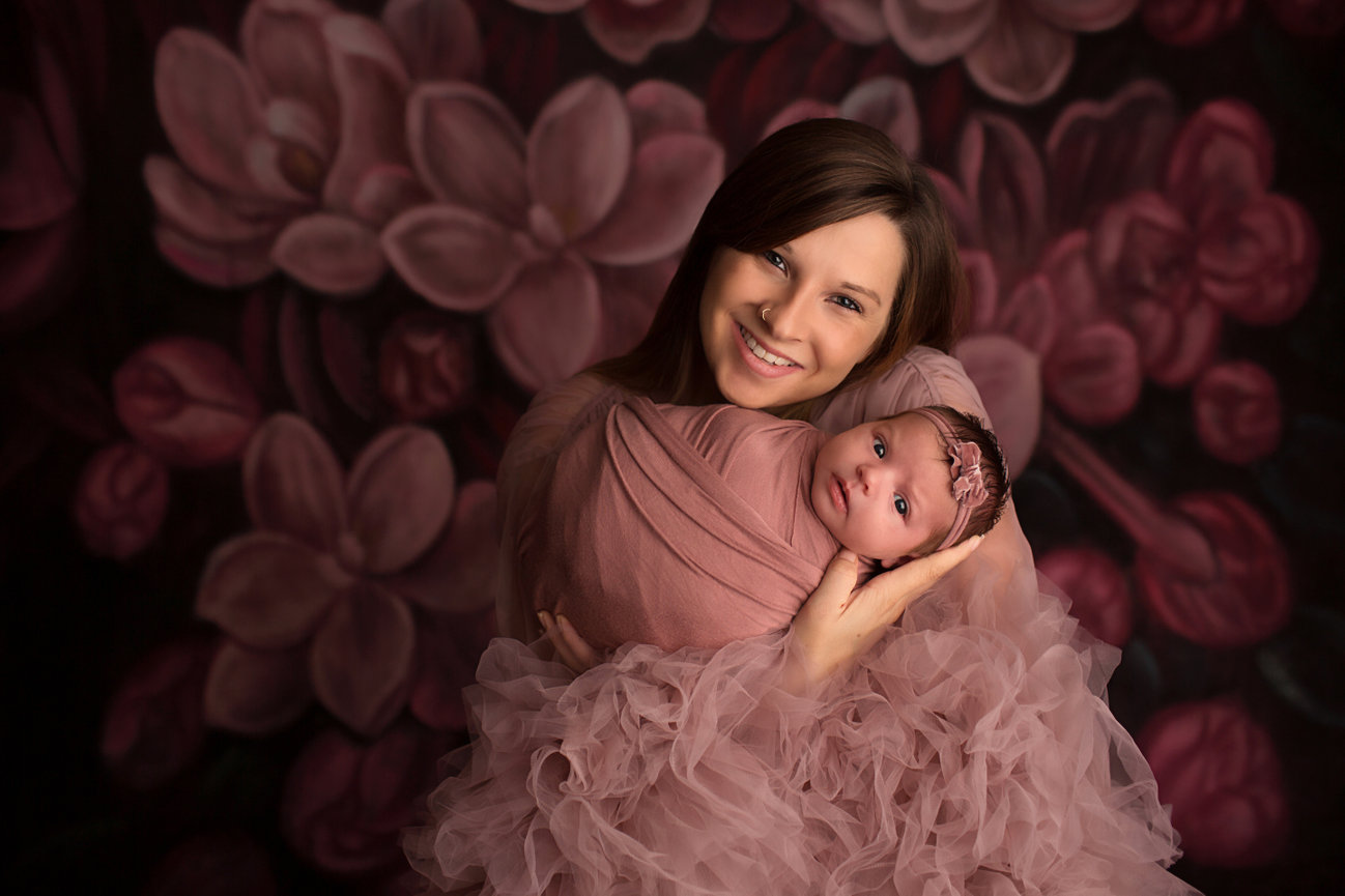 Newborn baby girl wrapped in mauve photographed with her mom holding her in front of beautiful floral backdrop during photoshoot by Kansas City newborn photographer Dreamworks Photography