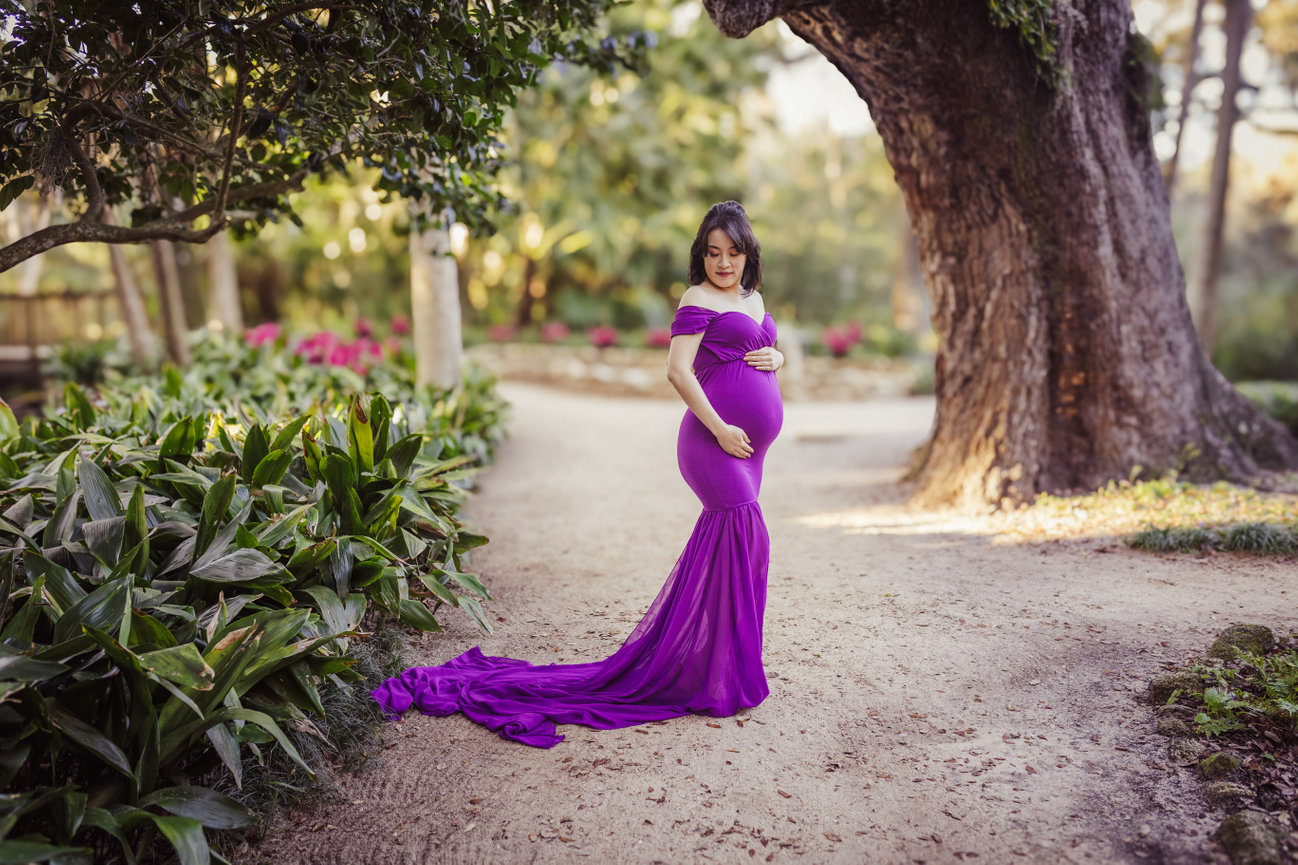 Pregnant woman in a flowing purple dress standing on a garden path surrounded by lush greenery and vibrant flowers at Washington Oaks Park during a maternity photography session with Catherine Whitney Photography in Jacksonville Florida