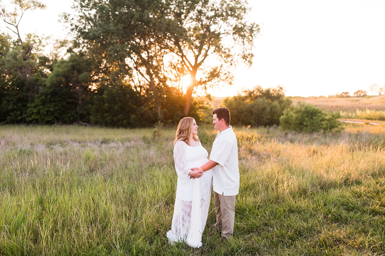 Couple in a field at sunset, standing together and holding hands, with trees and a grassy landscape in the background.