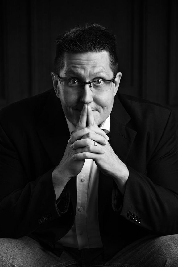 Man leaning forward on his knees with his fingers over his lips in a black and white headshot.