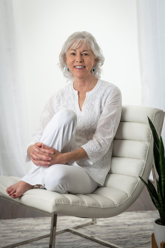 Smiling woman with gray hair in a white embroidered top and pants, seated on a modern chair against a white backdrop.