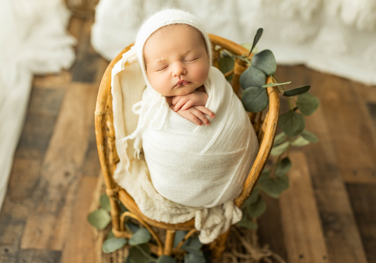 Newborn baby swaddled in white blanket sitting up in wicker chair