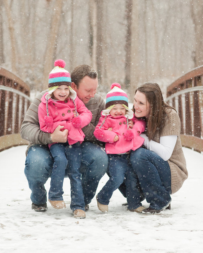 Family of four in colorful winter clothes, crouching and smiling on a snowy bridge, with snow gently falling around them.