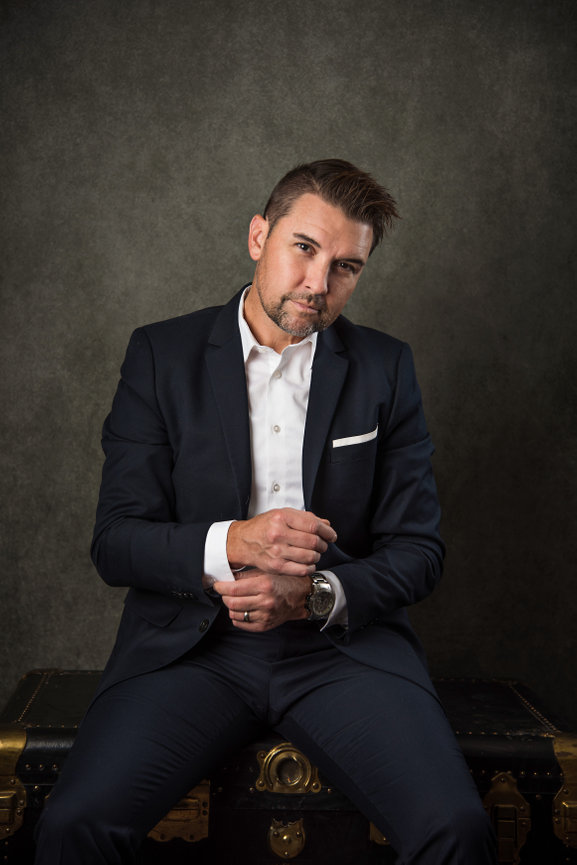 Man in a dark suit and white shirt sits on a vintage trunk, looking confidently at the camera.