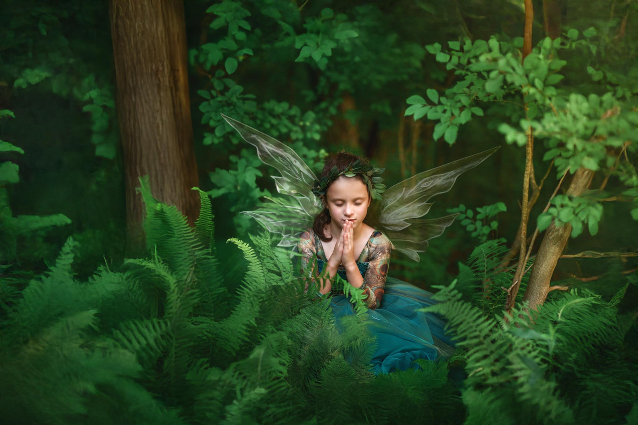 Fairy in a green gown praying while kneeling behind ferns on forest path near Pittsburgh, PA during a whimsical children’s session