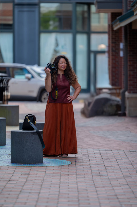 Jo Hayes Images holds a camera in a city street, wearing a maroon top and orange skirt, with a white car in the background on location for a photoshoot in downton main street