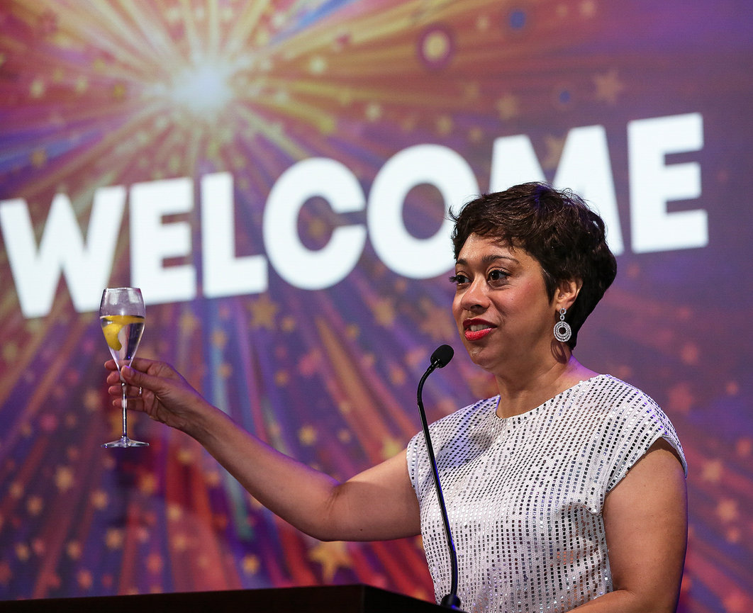 A woman in a white dress raises a champagne glass on stage with WELCOME in the background.