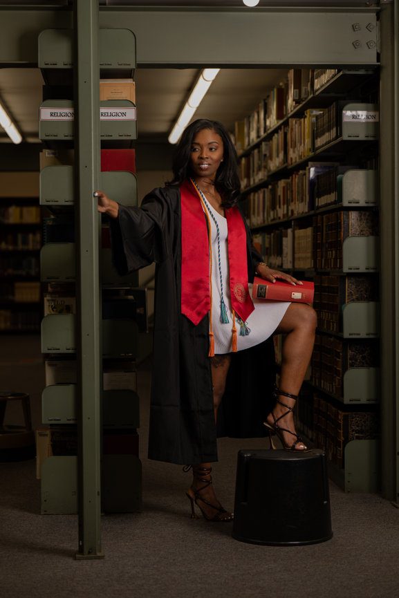Graduate in cap and gown poses confidently in a library, holding a diploma, surrounded by bookshelves captured by Jo Hayes Images Charlotte Portrait Graduate Photographer