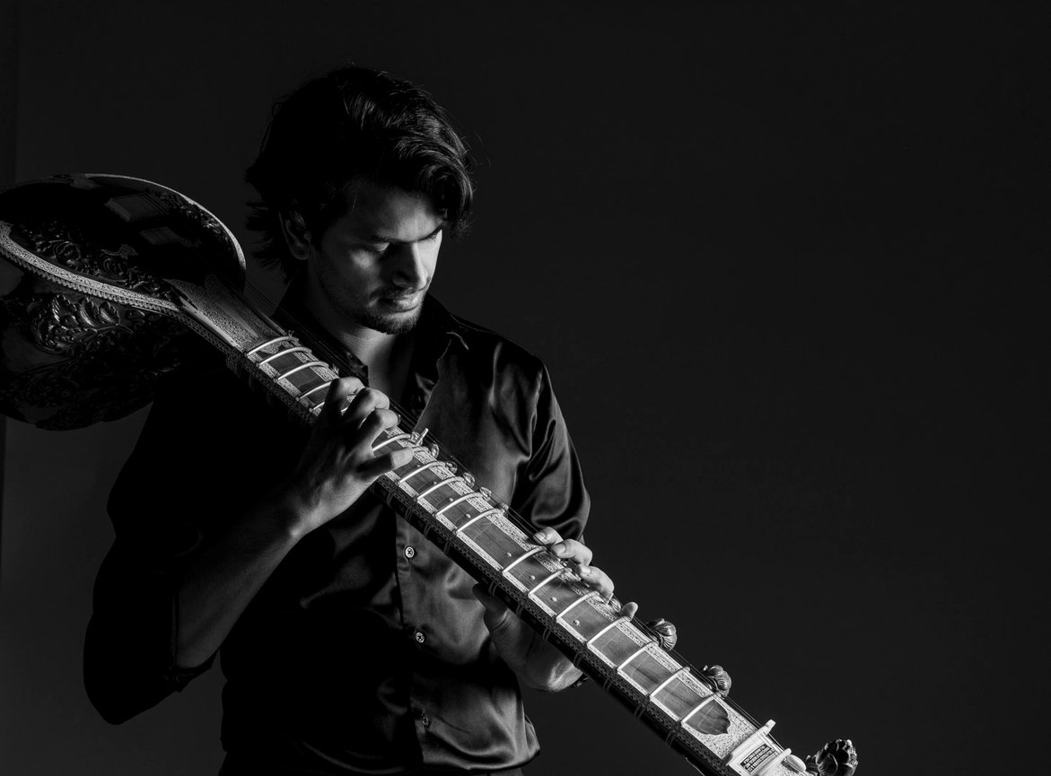 Black and white portrait of a man holding a sitar across his shoulder against a dark background with dramatic lighting