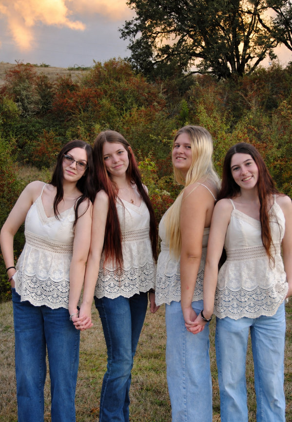 Four women standing together outdoors at sunset in jeans and white tops with trees in the background