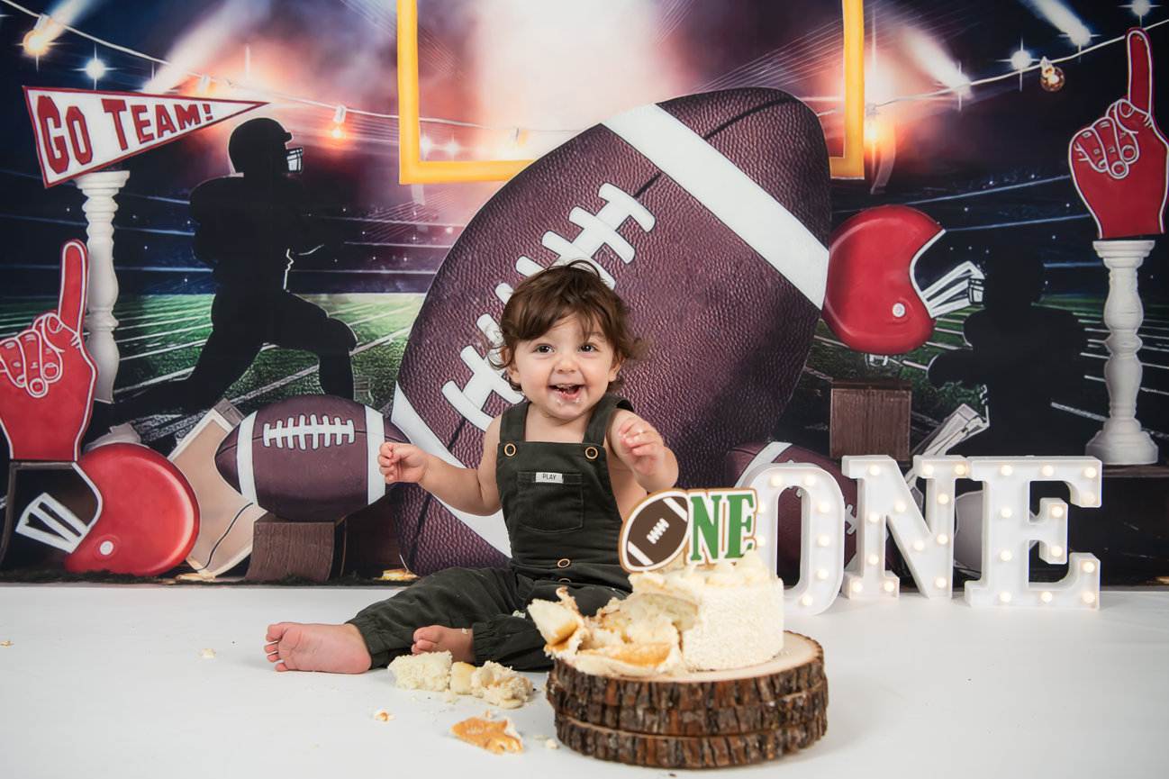 A joyful child sits on a backdrop of sports gear, surrounded by a cake, celebrating a first birthday.