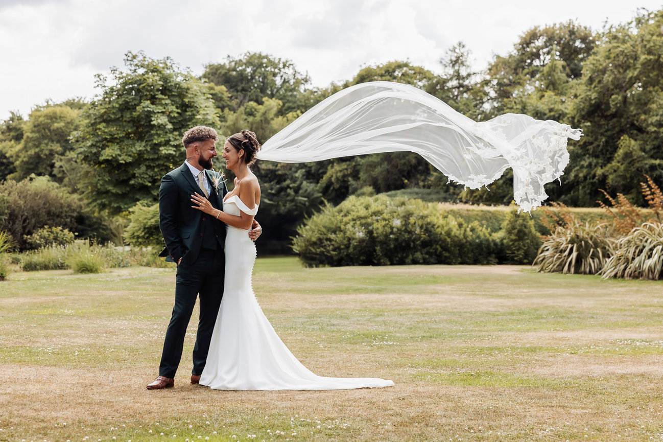 Bride and groom embracing outdoors, with the bride's veil blowing in the wind