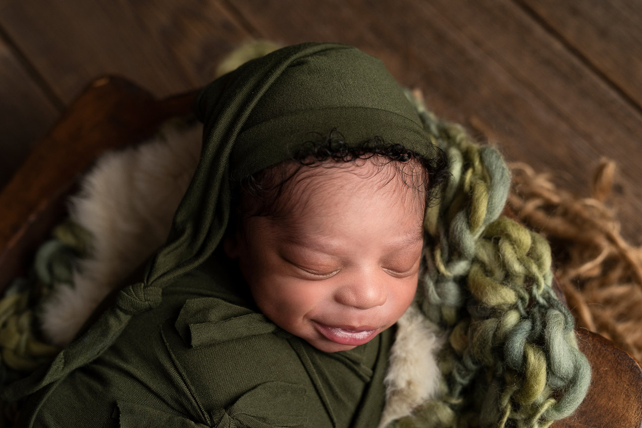 Newborn baby sleeps wrapped in a green swaddle with a matching cap. Nestled on a cozy, textured, blanket, the scene evokes warmth and comfort in Cary, Illinois, with its quaint and serene charm as the wooden backdrop completes this tranquil setting.