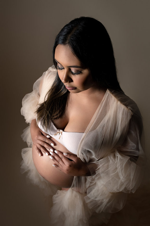 Pregnant woman in a sheer, flowing robe with her hands gently resting on her belly. She gazes down, creating a serene and intimate atmosphere.