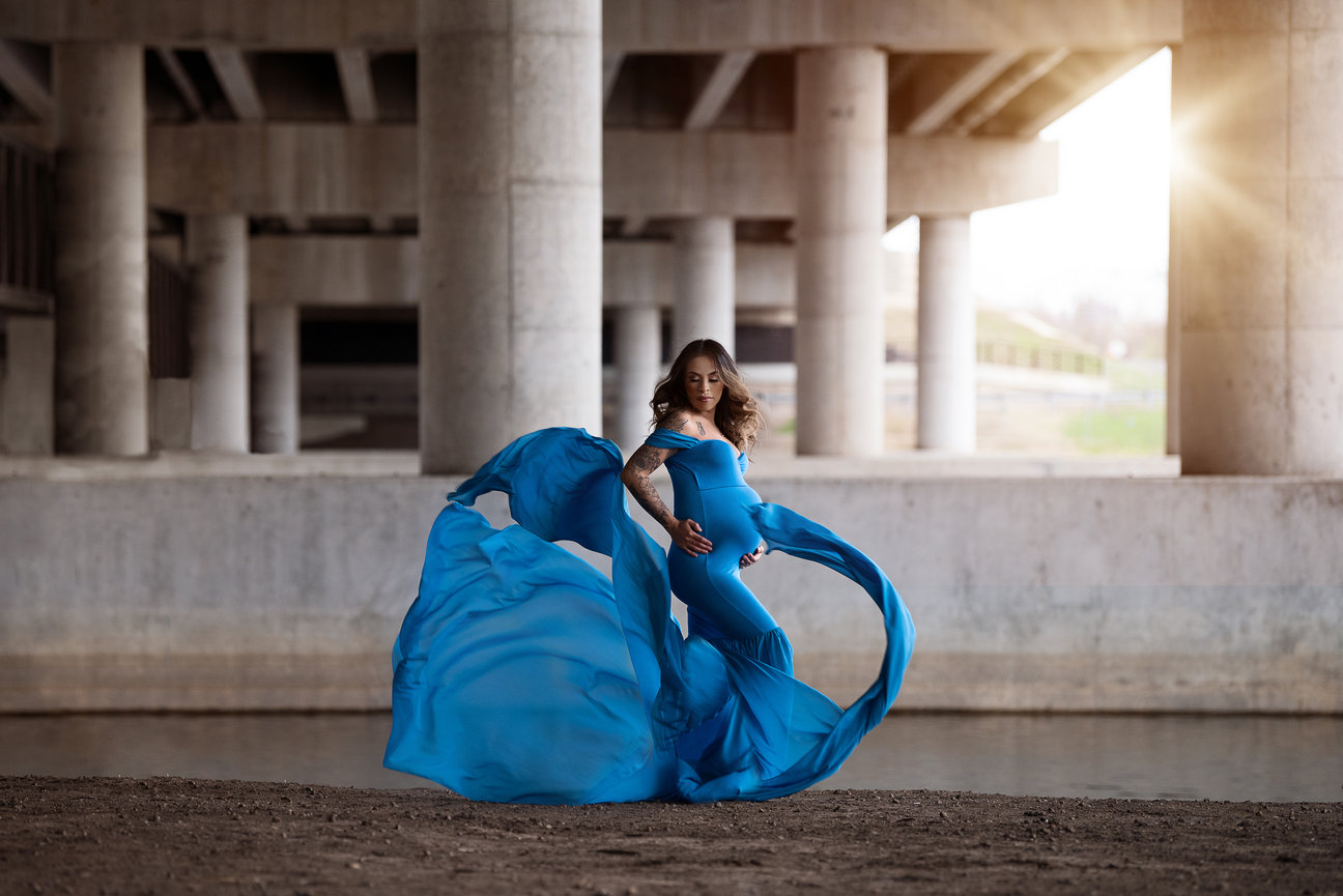 A woman in a flowing blue dress poses gracefully under a concrete bridge, capturing the essence of a serene maternity shoot. The dress billows dramatically around her, and the sunlight filters through the columns, casting a warm glow.