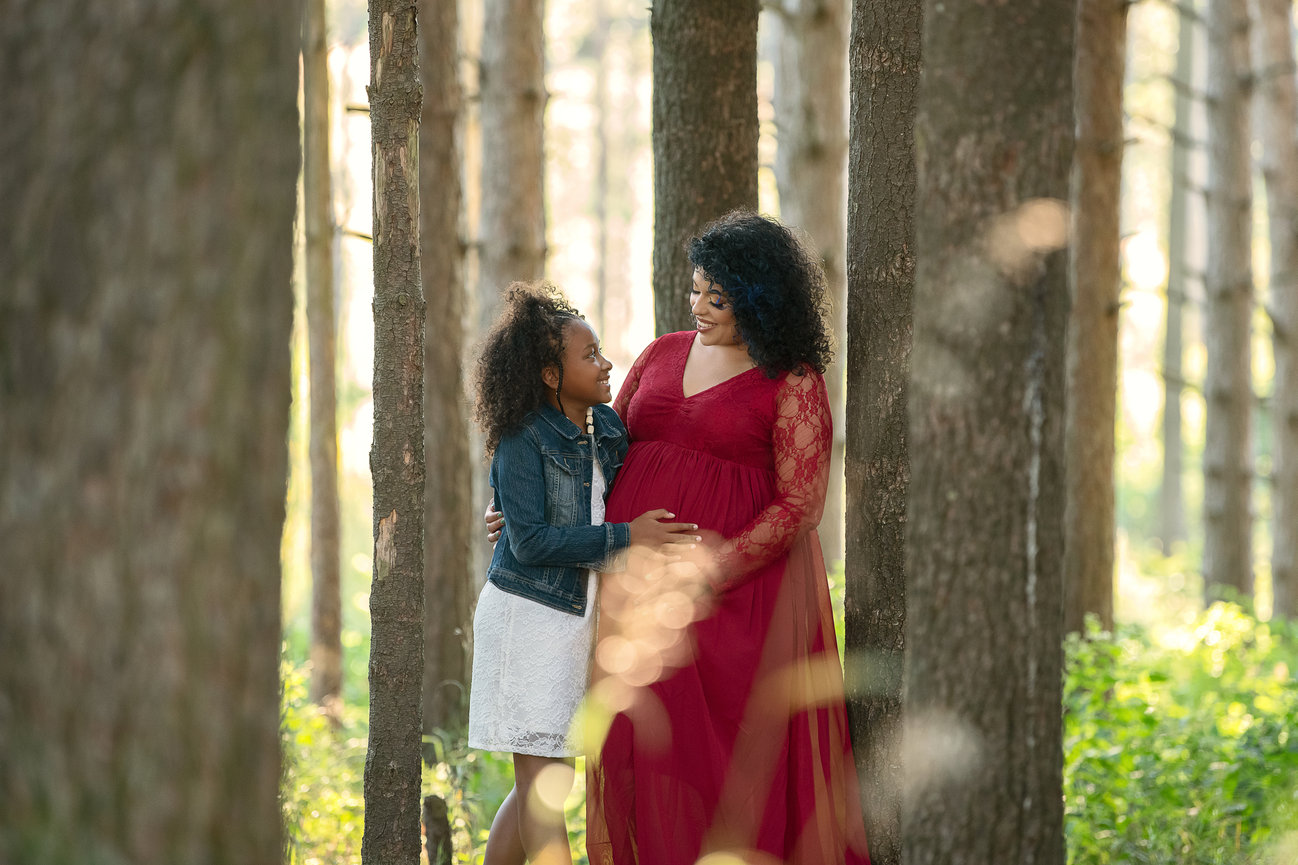 A woman in a long red dress and a young girl in a white dress and denim jacket stand closely together, embracing and smiling, in a sunlit forest with tall trees.