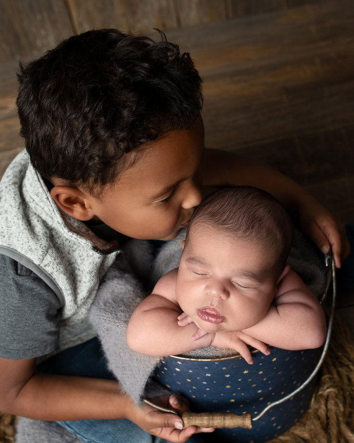 In Huntley, a young child gently kisses a sleeping baby on the forehead, snuggled in a textured basket with a soft blanket. The scene is set against a warm, wood-like floor, capturing the essence of small-town charm.