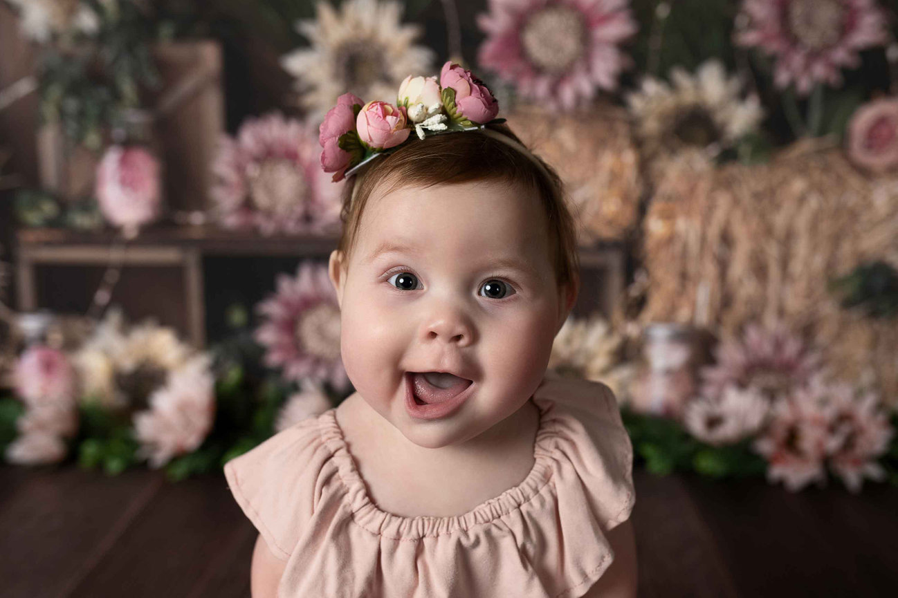 A joyful twelve-month-old baby with brown hair is wearing a pink dress and a floral headband. With an open mouth, the baby radiates happiness. The background is adorned with hay bales and pink flowers.